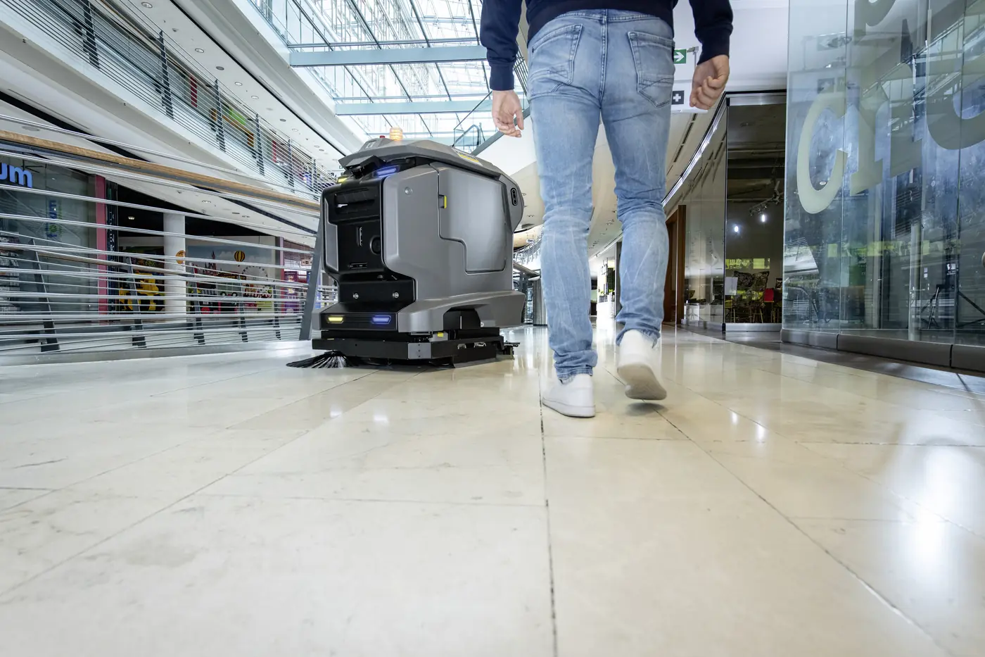 Passant geht in einem Einkaufszentrum an einem reinigenden KIRA Scheuersaugroboter vorbei